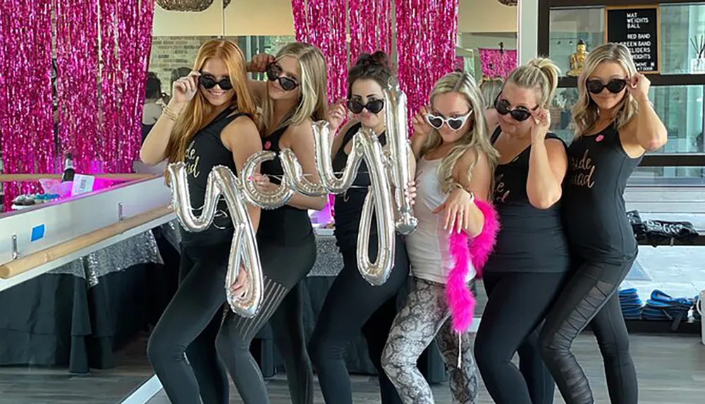 A group of women wearing matching Bride Squad tank tops and sunglasses are playfully posing together with metallic balloon letters at what appears to be a bachelorette party