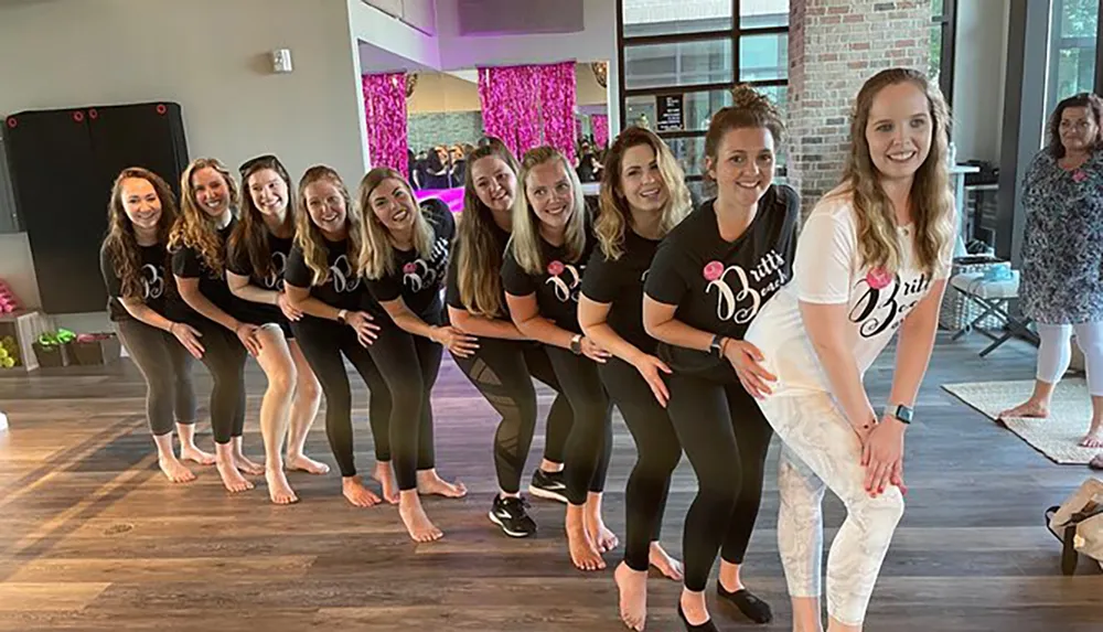 The image shows a group of smiling women posing playfully in matching t-shirts in what appears to be a dance or exercise studio with one woman standing out in a white t-shirt and pants