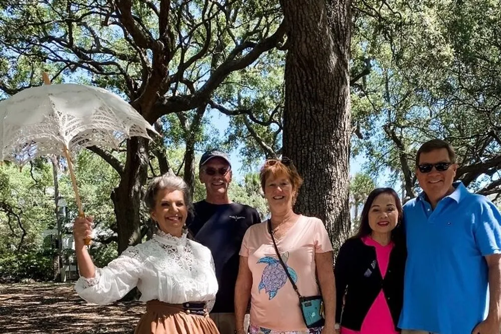 Five people are smiling for a photo outdoors with one person dressed in a historical costume holding a lace parasol under large leafy trees
