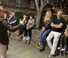 A person is gesturing enthusiastically while speaking to a group of smiling people casually seated on a brick ledge outdoors at night