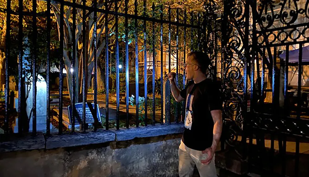 A person is standing at night by a wrought-iron fence that encloses a well-lit park or garden area