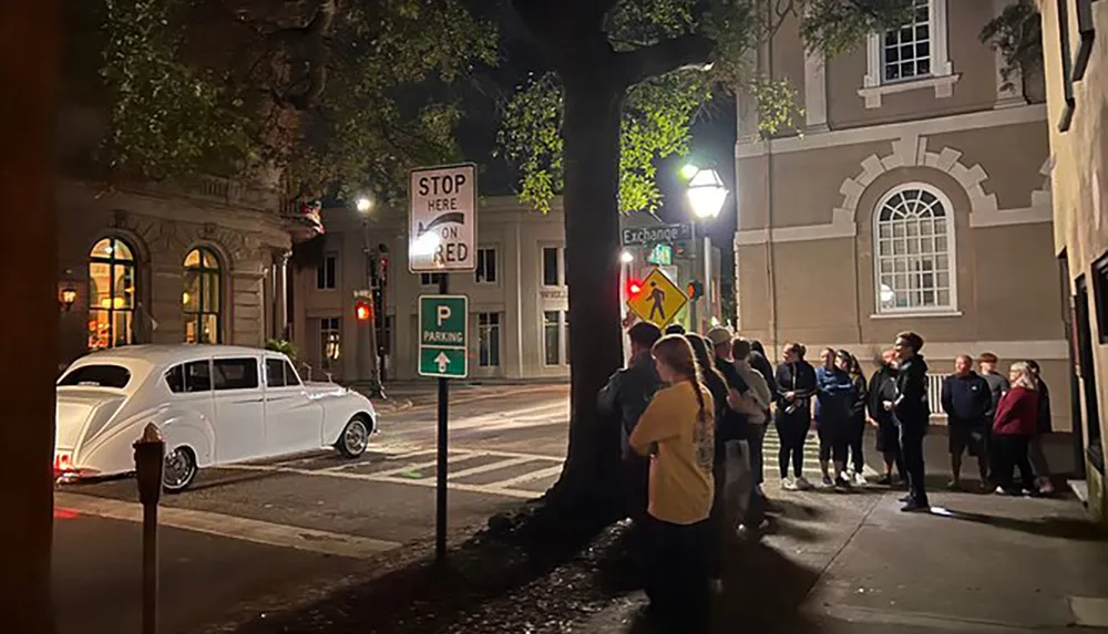 A group of people is gathered on a city sidewalk at night with some looking upwards while a vintage white car is parked near a stop sign across the street