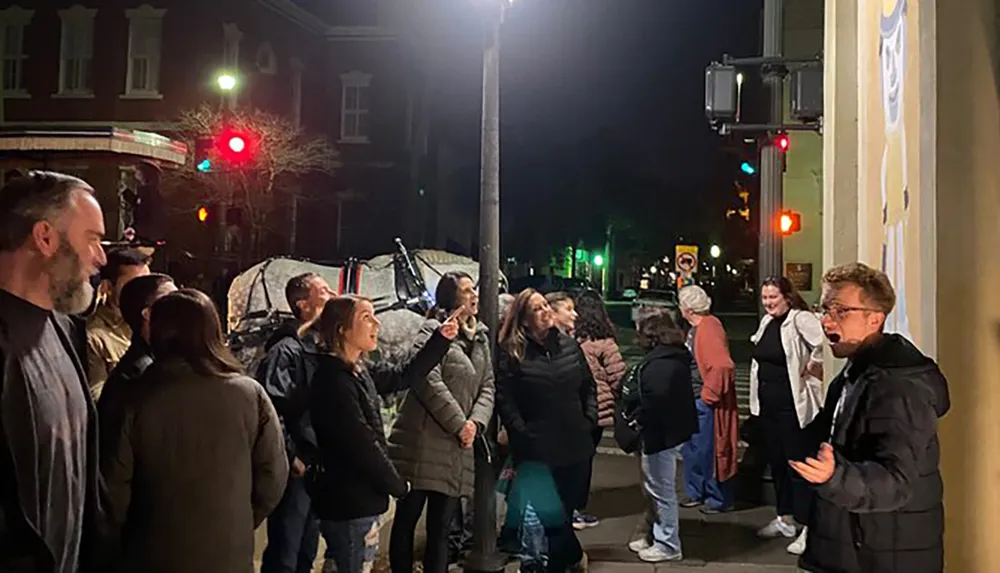 A group of people is gathered at night on a city street focusing their attention on something above them out of the cameras view