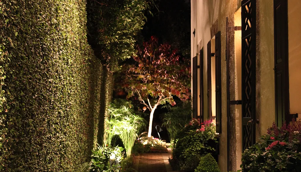 This image depicts a warmly lit garden pathway at night bordered by lush greenery and a building with elegant windows and shutters