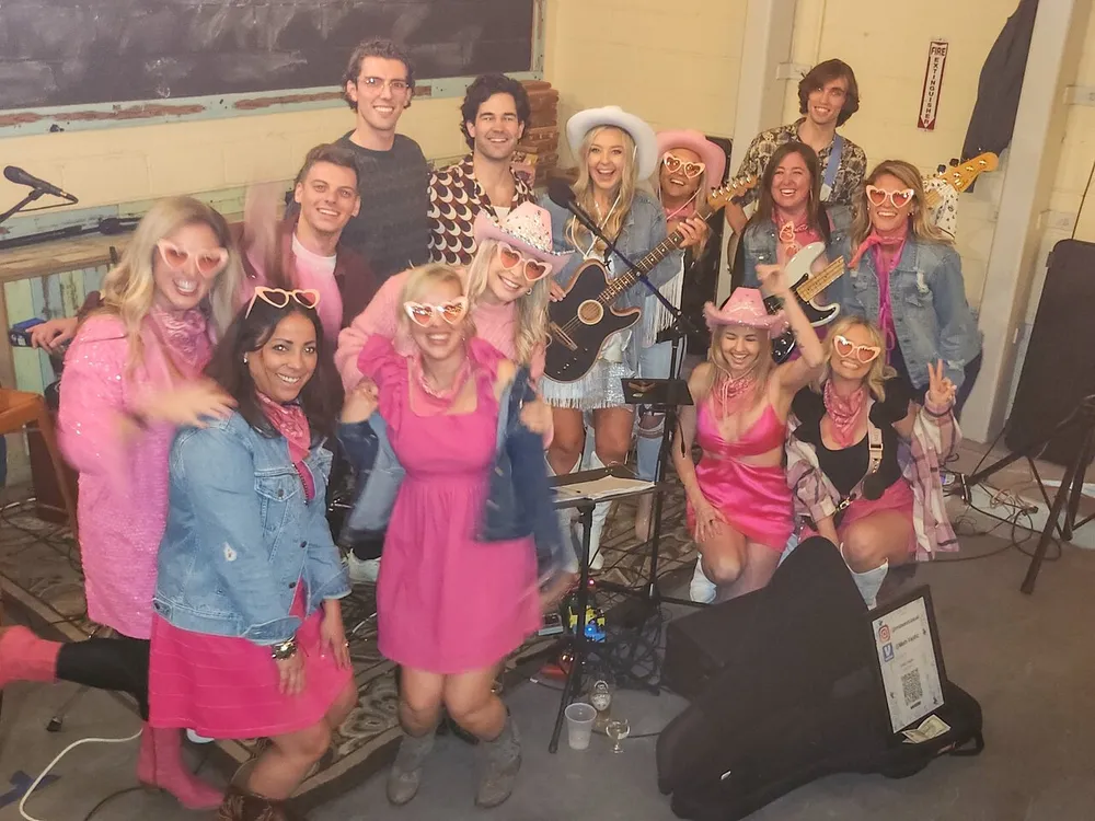 A group of cheerful people is posing for a photo with musical instruments wearing pink-themed outfits some with heart-shaped glasses and cowboy hats suggesting a festive or themed event