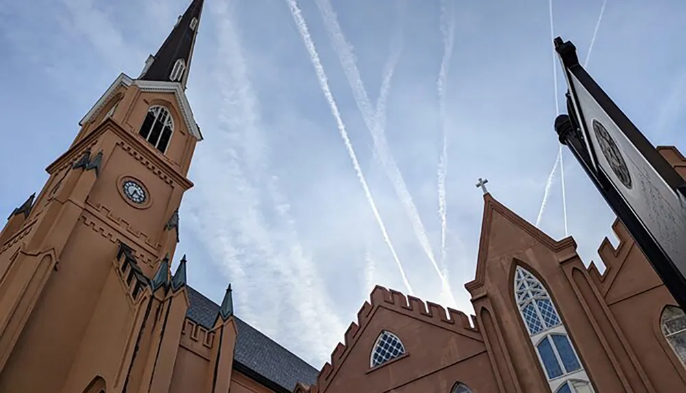 The photo captures a salmon-colored church with a tall spire set against a blue sky crisscrossed by the linear contrails of airplanes