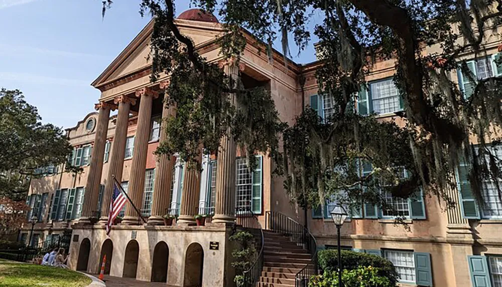 The image shows a stately historic building with a domed roof and large pillars adorned with Spanish moss hanging from trees and an American flag out front