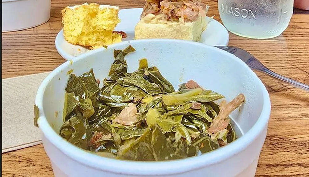 A bowl of collard greens is accompanied by cornbread and a biscuit with meat on top evoking a traditional Southern meal with a glass of water on the side