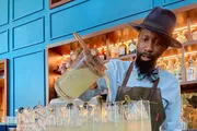 A bartender in a hat and apron is pouring a cocktail from a mixing jug into glasses lined up on a bar counter.