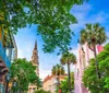 The image shows a picturesque street lined with colorful buildings a church spire palm trees and lush greenery under a bright blue sky