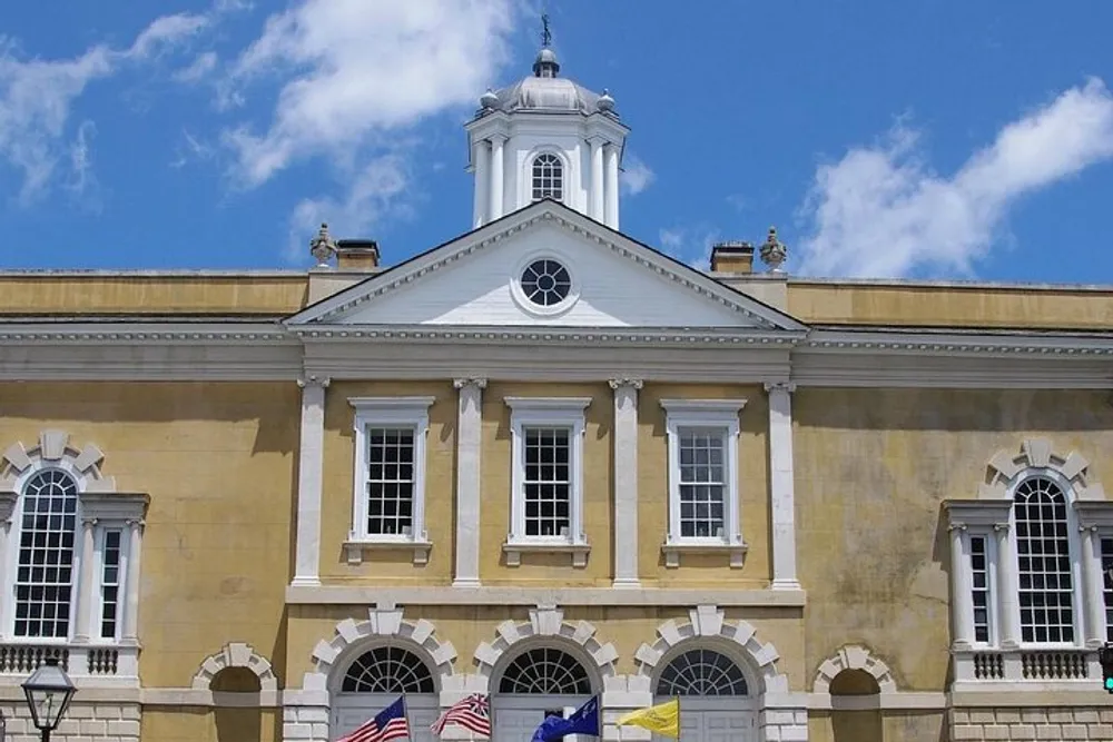 The image shows a grand neoclassical building with a central clock tower numerous windows and multiple flags including the United States flag under a clear blue sky