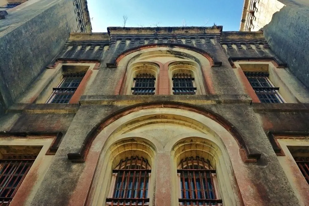The image shows the weathered facade of an old building with arched windows that are secured with bars looking up at a clear blue sky