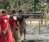 Two individuals dressed in historical clothing are aiming long rifles in an outdoor setting that suggests a historical reenactment or demonstration