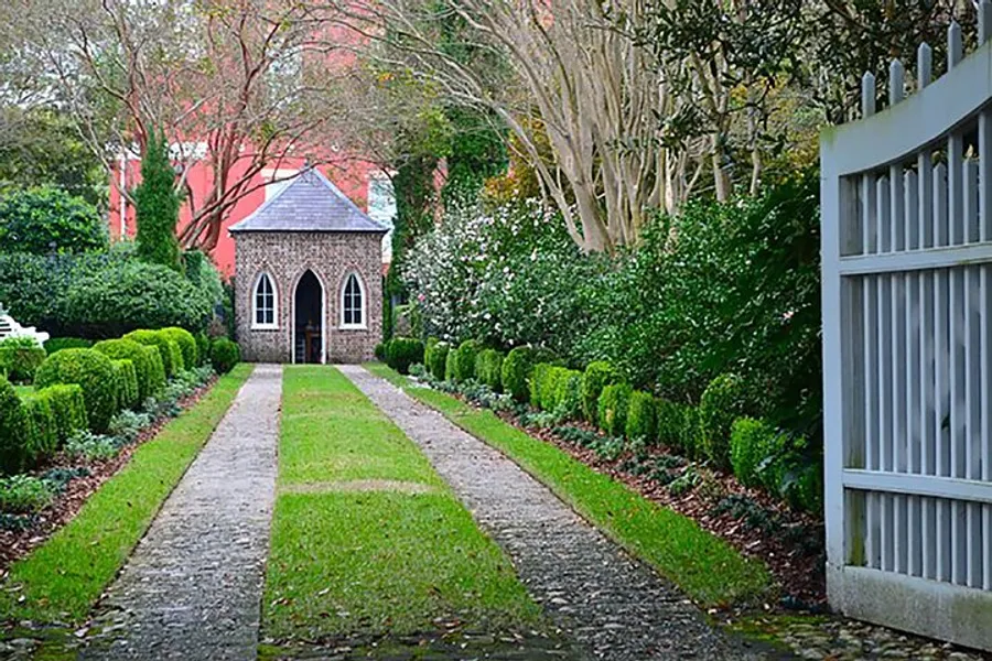 A quaint small stone chapel is nestled at the end of a neatly trimmed hedged pathway, flanked by lush greenery and a welcoming open gate.