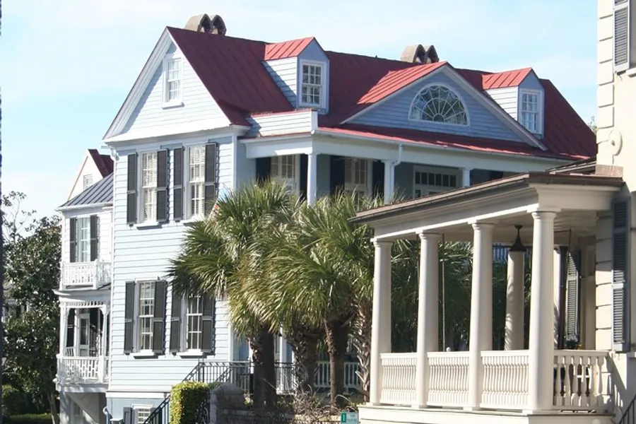 The image shows a traditional two-story white house with a red roof, featuring a front porch with columns, shuttered windows, and a prominent dormer window, set against a backdrop of clear skies and framed by palm trees.