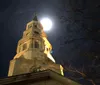 The image shows the tower of a church at night with the full moon appearing directly behind the spire creating a dramatic and picturesque scene