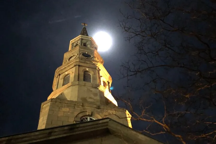 The image shows the tower of a church at night, with the full moon appearing directly behind the spire, creating a dramatic and picturesque scene.