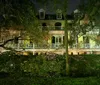 The image shows a grand multi-story house with balconies illuminated at night by exterior lights surrounded by lush greenery and trees