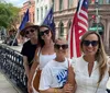 Four smiling women pose together on a sunny city street corner flanked by American flags and against a backdrop of historic buildings