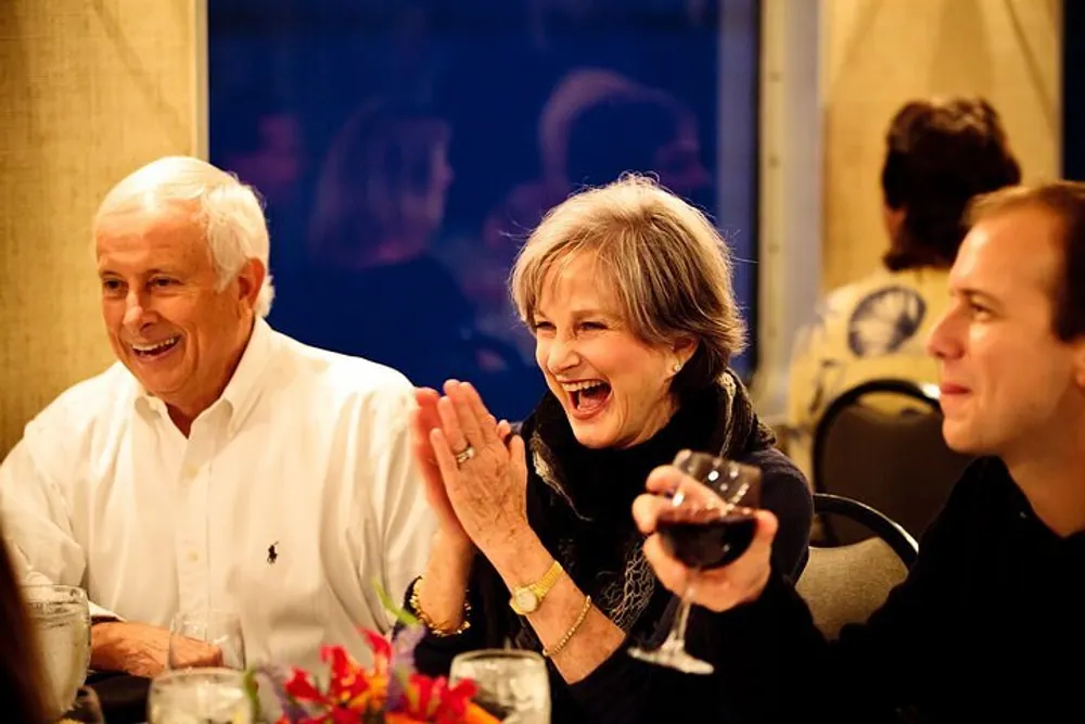 A group of people are enjoying a lively conversation at a dinner table with one woman laughing cheerfully while holding a glass of red wine