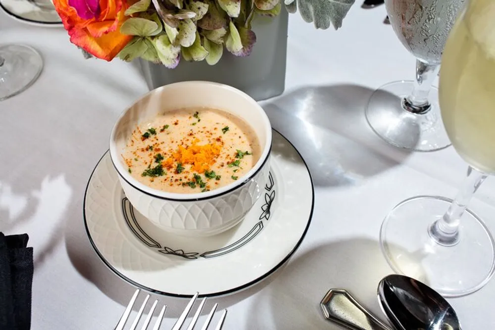 An elegant table setting featuring a bowl of creamy soup garnished with herbs and spices flanked by a glass of wine and silverware on a white linen tablecloth