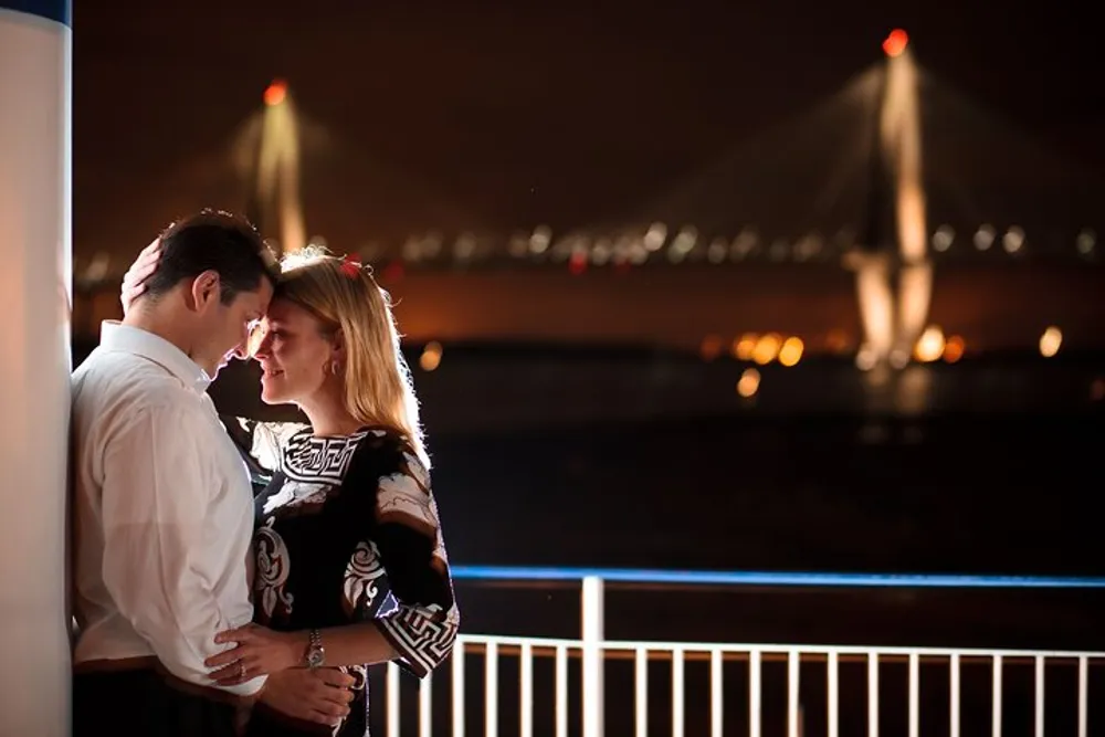 A couple shares an intimate moment on a balcony with a softly lit suspension bridge in the background at night