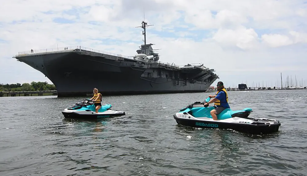 Two individuals on personal watercrafts are floating near a large aircraft carrier docked at a port