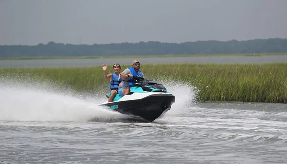 Two people in life jackets are enjoying a fast ride on a jet ski across a body of water with green reeds in the background