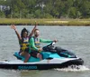 Two people are smiling and waving as they ride a Sea-Doo jet ski on a sunny day with calm water and greenery in the background