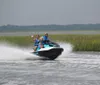 Two people are smiling and waving as they ride a Sea-Doo jet ski on a sunny day with calm water and greenery in the background