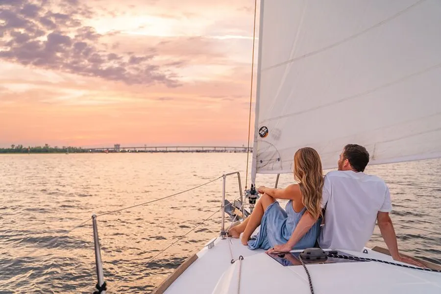 A couple is sitting on the deck of a sailboat, enjoying a serene sunset over the water.