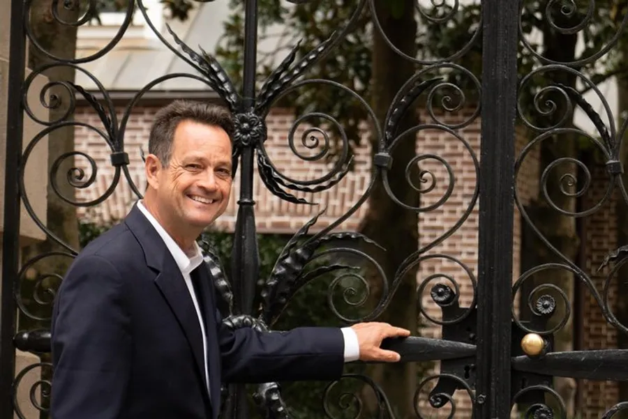 A person is smiling at the camera while standing at an ornate wrought-iron gate with a brick building in the background.