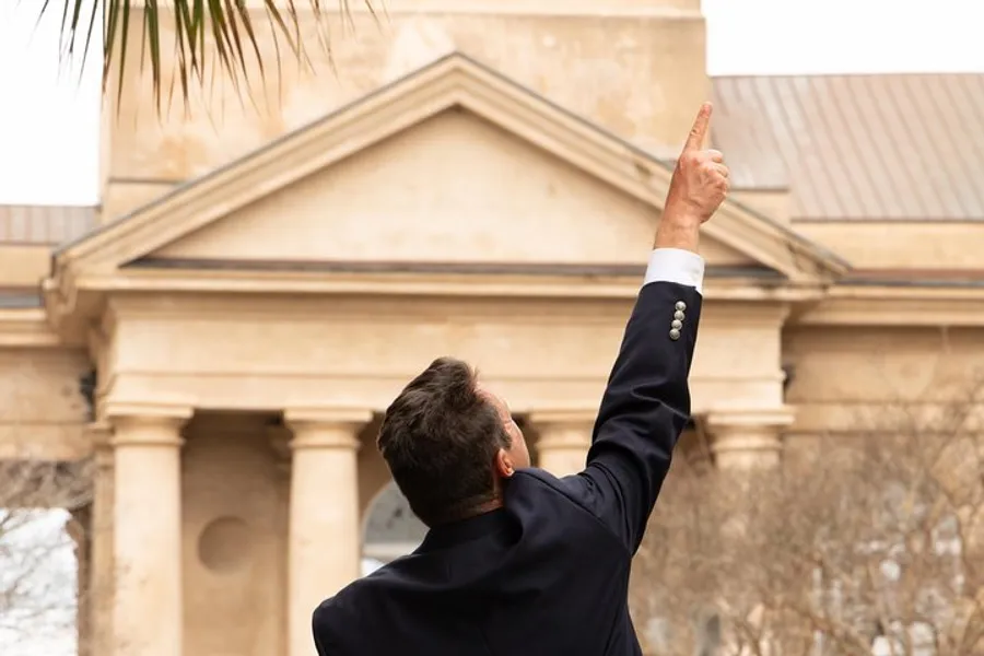 A person in a dark suit is seen from behind, pointing upwards with their index finger against a background featuring classical architecture.