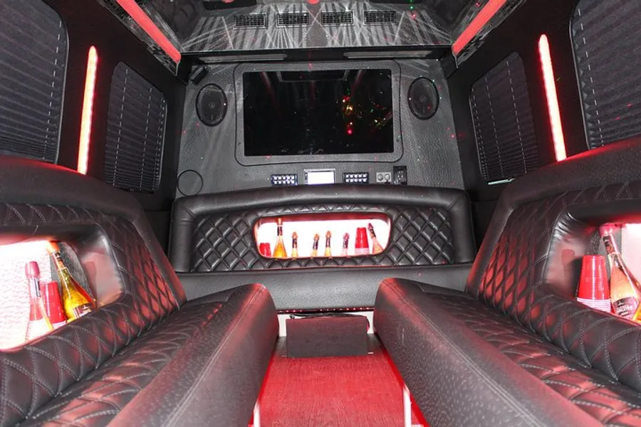 The image shows an opulent and modern limousine interior featuring red LED lighting, black quilted leather seats, a spacious cabin, and a central bar area with a large screen on the front wall.
