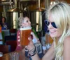 A smiling person wearing sunglasses is holding up a glass of beer in a brewery with other people and brewing equipment in the background