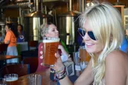 A smiling person wearing sunglasses is holding up a glass of beer in a brewery with other people and brewing equipment in the background.