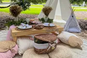 The image shows a cozy outdoor picnic setup with a low table, comfortable cushions, a basket, and decorative elements, including a sign that says 