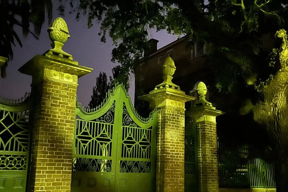 An ornate gate with intricate patterns and statues atop its pillars is cast in a mysterious green light against the night sky