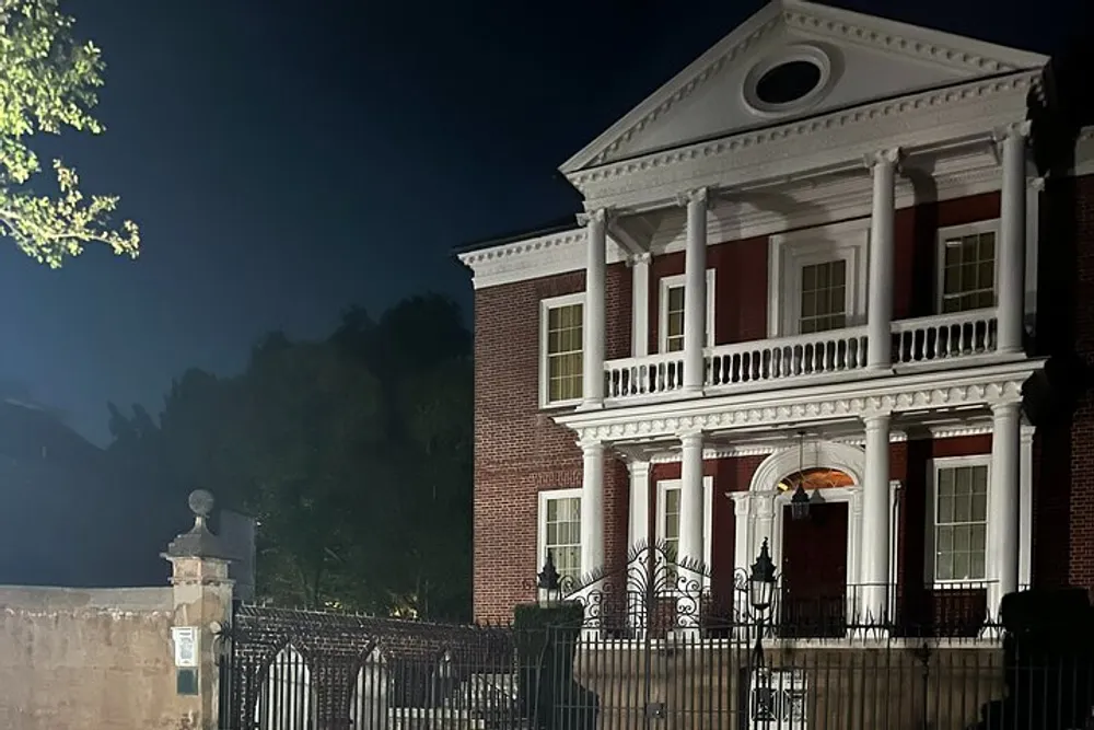The image displays a stately two-story red brick building with white columns and a balcony at night accentuated by exterior lighting with a wrought iron fence in the foreground