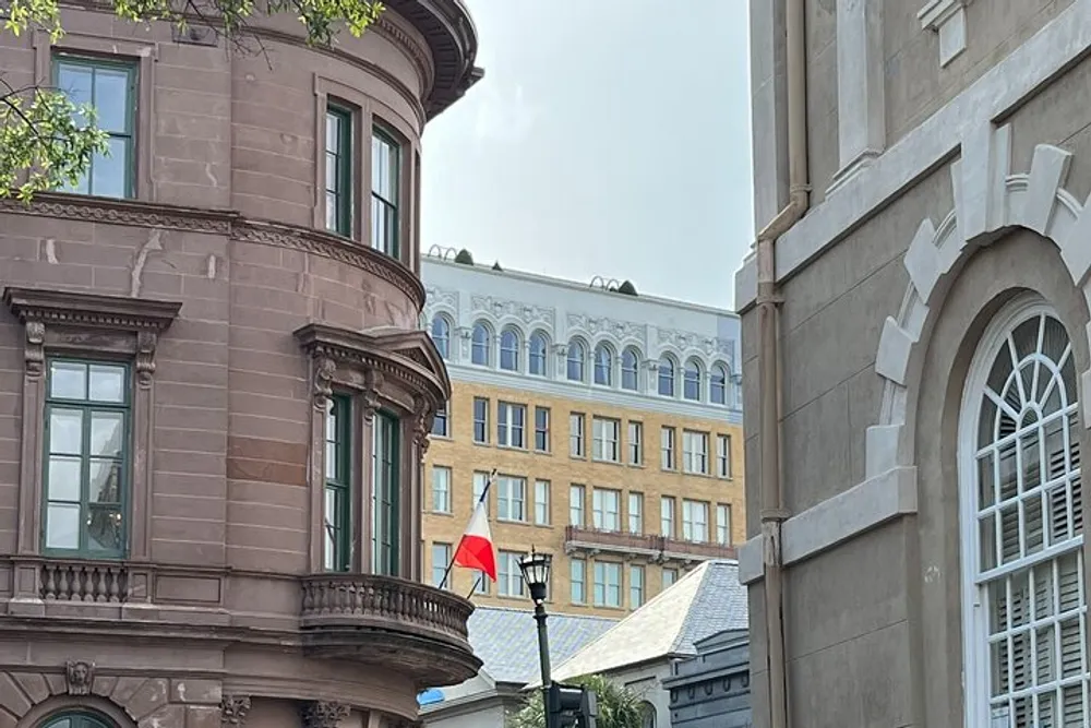 The image shows a street corner with historic architecture featuring buildings with ornate detailing and a red flag hanging from a rounded balcony set under a cloudy sky