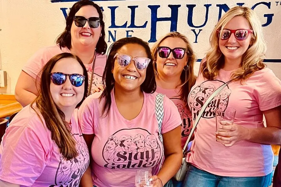 A group of five women are smiling for the camera, wearing matching pink t-shirts and sunglasses indoors, likely participating in a social event.