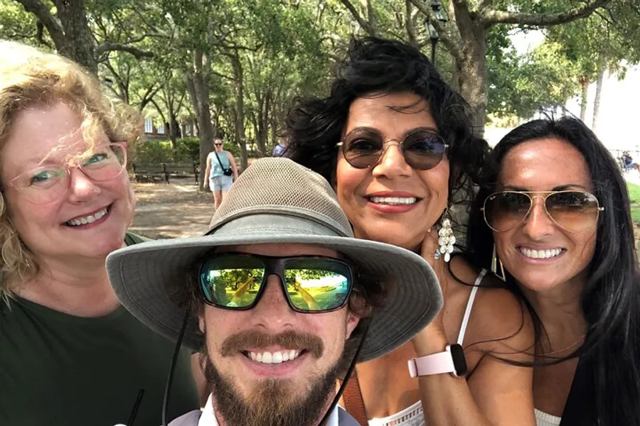 Four smiling people are taking a selfie in a sunny, tree-filled outdoor setting.