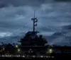 An aircraft carrier is illuminated by string lights and deck lighting under a dusky sky creating a dramatic and imposing silhouette