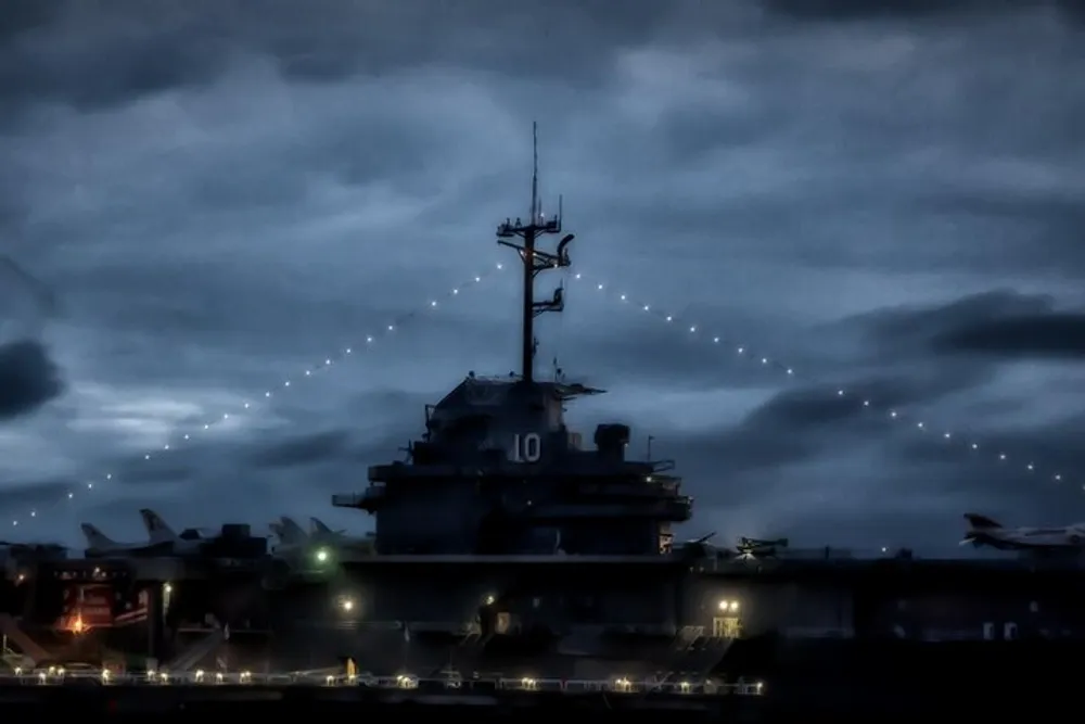 An aircraft carrier is illuminated by string lights and deck lighting under a dusky sky creating a dramatic and imposing silhouette