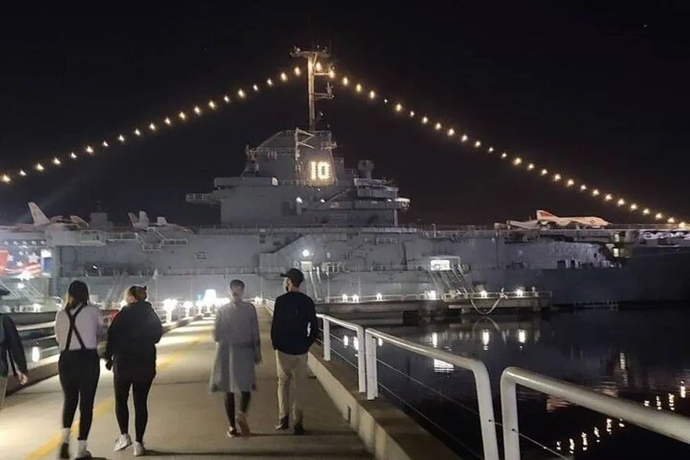A group of people walks along a pier at night illuminated by string lights with a large aircraft carrier moored on the right