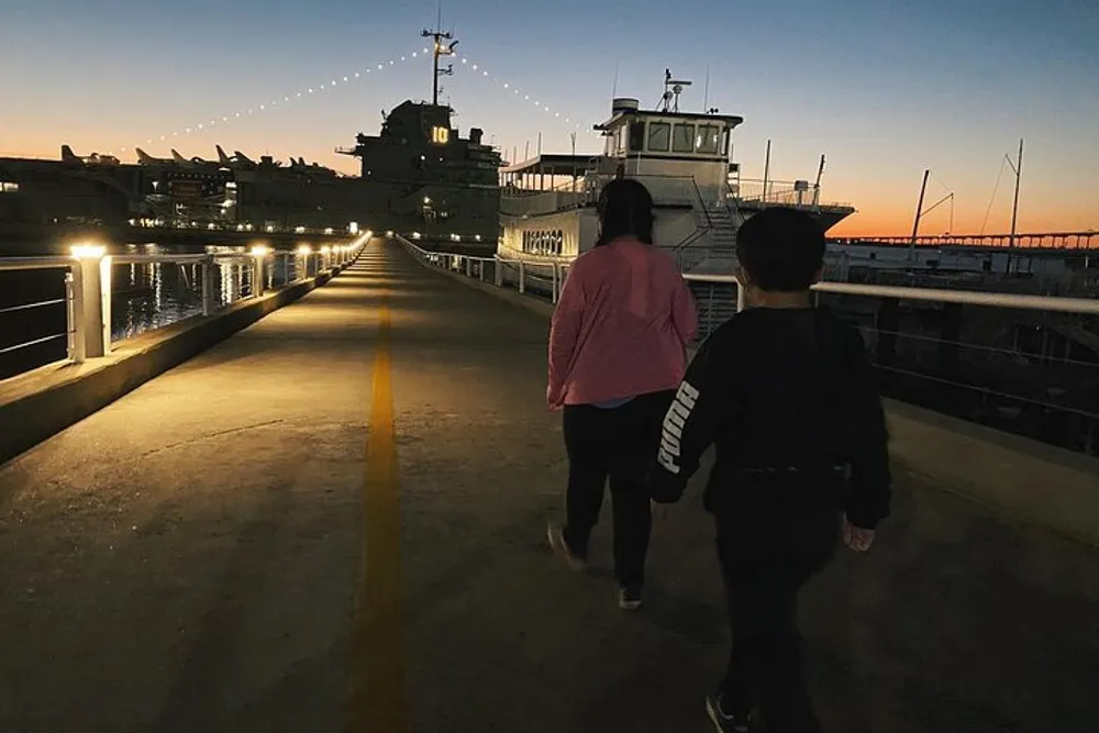 Two people are walking along a dock at dusk with lights illuminated along the handrails and a ship moored to the right