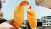 Two hands are holding glasses filled with a bubbly orange beverage, garnished with orange slices, in a sunny outdoor setting.