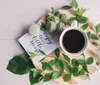 A cup of coffee surrounded by green macarons fresh leaves and a note saying enjoy the little things rests on a wooden surface