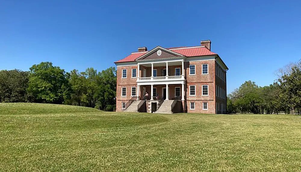 The image shows a large two-story brick house with a red roof and white columns nestled in a grassy landscape under a clear blue sky
