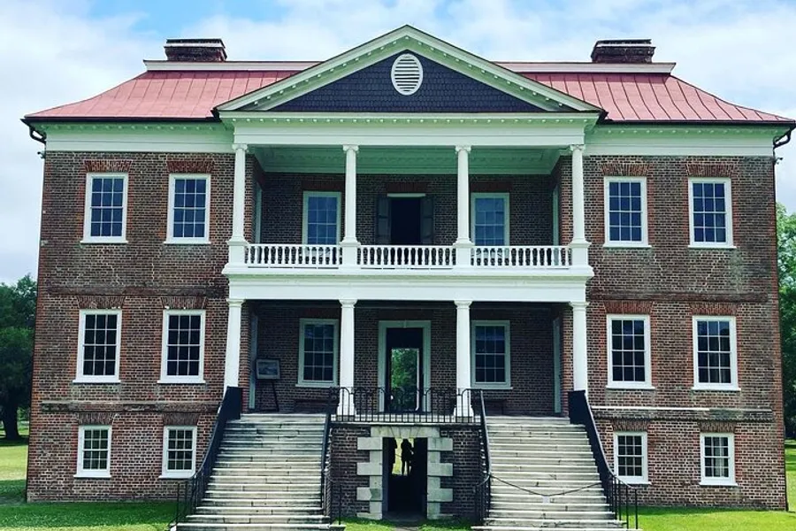 The image shows a symmetrical two-story brick house with a red roof, featuring a central pediment with a circular vent, a two-level porch with columns, and twin staircases leading to the entrance.